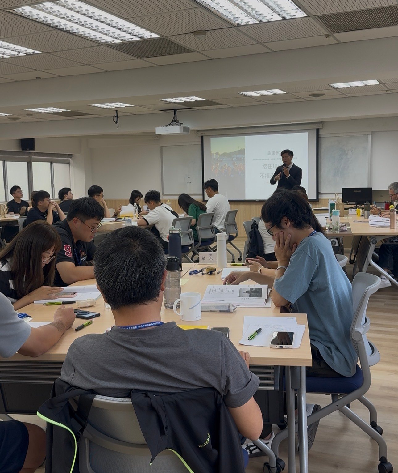 台電第一線服務人員培訓現場,Hydra 夏承樟持麥克風講解,學員分桌就座並閱讀教材。